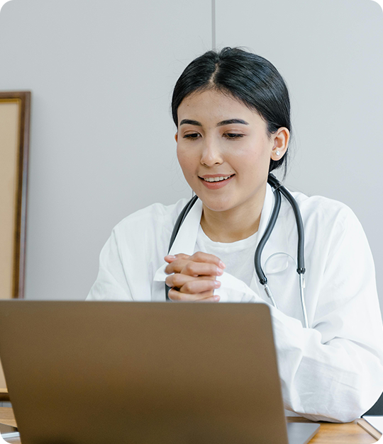 A woman in white lab coat sitting at table with laptop.